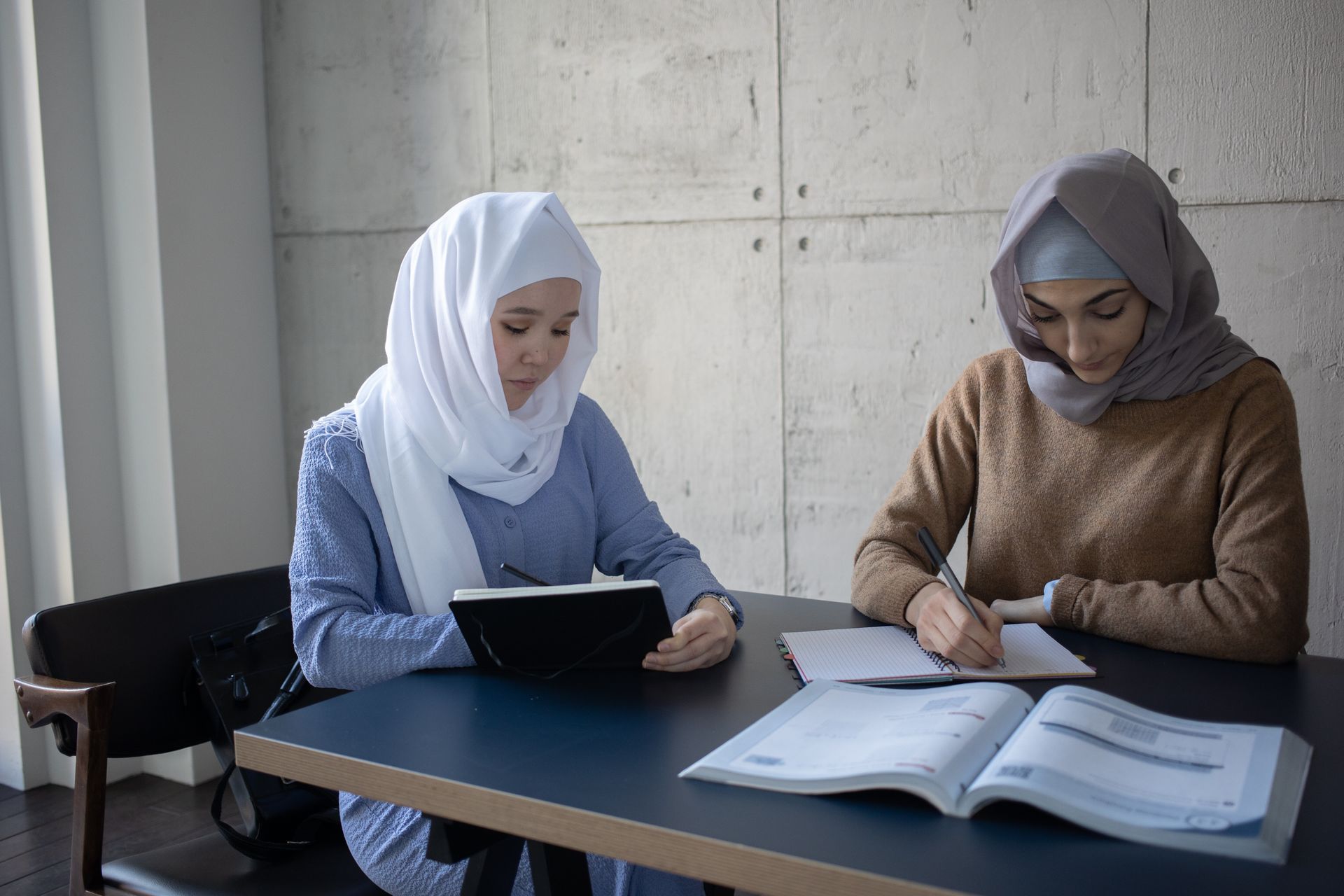 pexels-gabby-k-6282020-2 - Blog Muslim Marriage Services Two women wearing headscarves sit at a table studying: one using a tablet, the other writing in a notebook against a concrete wall backdrop.