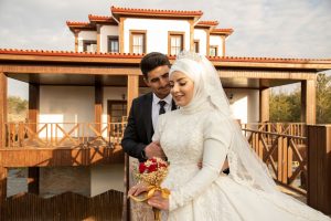 Bride in a white lace wedding dress and veil, holding a bouquet, with a groom embracing from behind beside a wooden building.