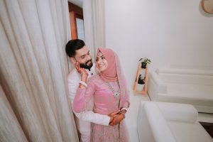 Bride in pink traditional dress and hijab hugs groom in a white suit beside beige curtains, henna on hands visible, indoor setting.