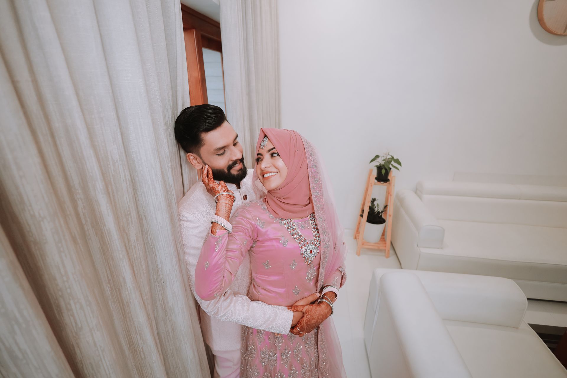 pexels-soulkidphotography-26990489-2 - Blog Muslim Marriage Services Bride in pink traditional dress and hijab hugs groom in a white suit beside beige curtains, henna on hands visible, indoor setting.