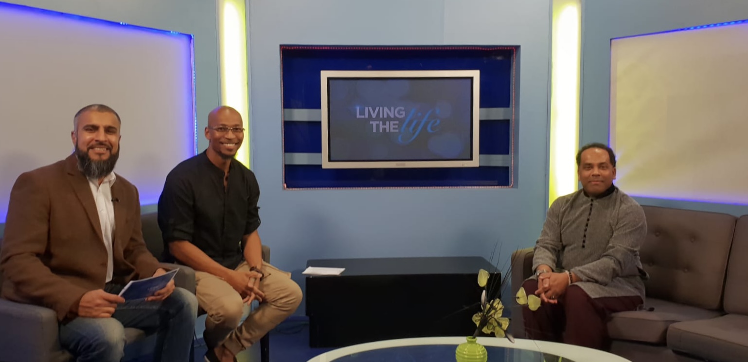 Three men sit on a TV talk-show set with a center screen reading 'Living the life'.