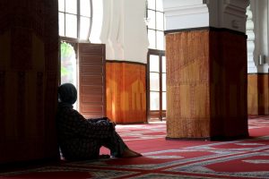 Woman wearing a headscarf sits on a red patterned carpet beside a large pillar inside a sunlit mosque.