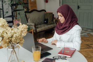 Woman in a burgundy hijab typing on a laptop at a table, with dried flowers and a glass of orange juice nearby in a cozy home setting, focused on work.