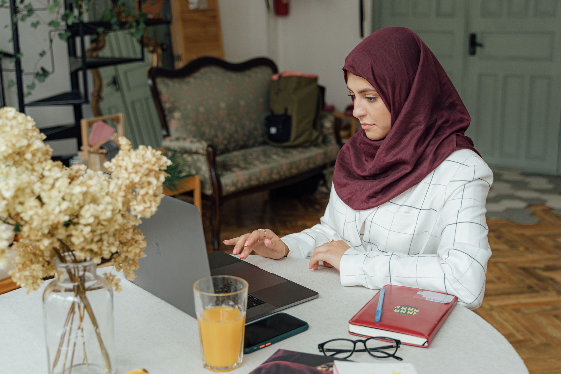 Woman in a burgundy hijab typing on a laptop at a table, with dried flowers and a glass of orange juice nearby in a cozy home setting, focused on work.