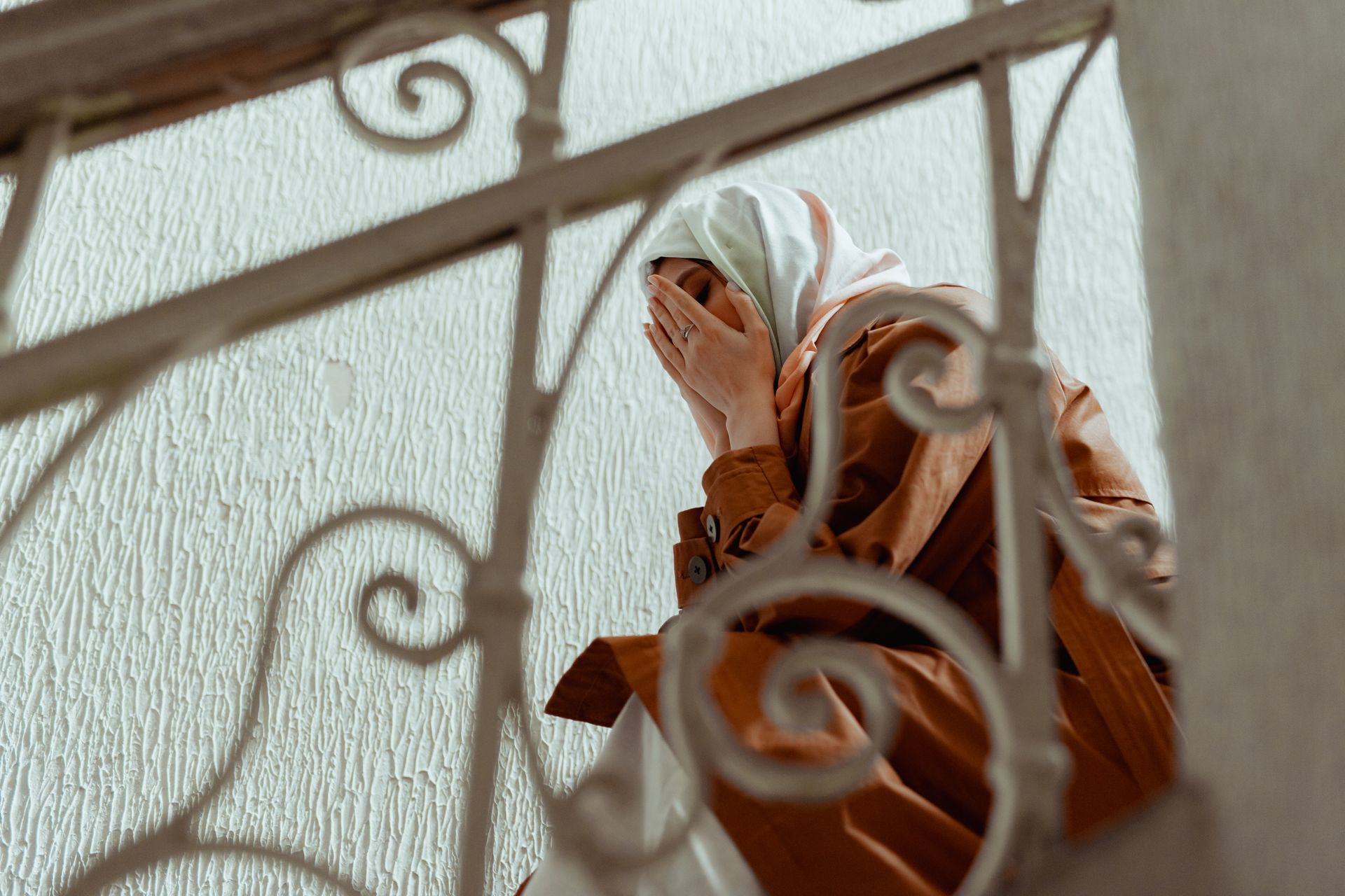 pexels-mikhail-nilov-7929089 - Blog Muslim Marriage Services Woman wearing a white headscarf and brown coat covers her face with her hands on a stairwell railing, conveying distress or sadness.