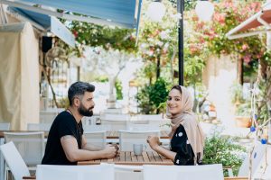 Two people seated at an outdoor cafe table, chatting and smiling, with flowers and plants in the bright background.
