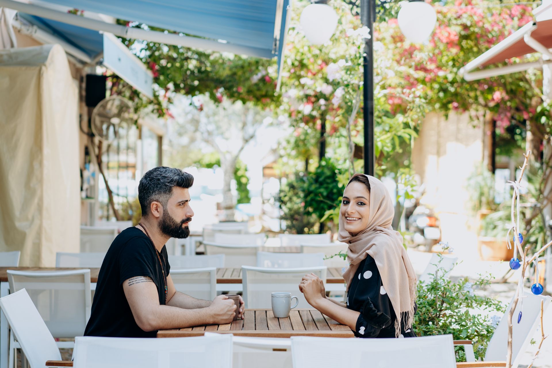 Two people seated at an outdoor cafe table, chatting and smiling, with flowers and plants in the bright background.