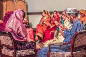 Front row: a man in a blue traditional outfit and cap praying, beside a woman in a pink sari, at a seated ceremony.