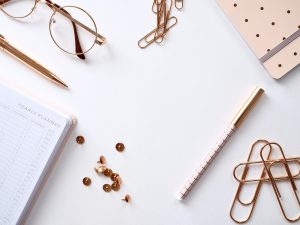 Flat lay of rose-gold stationery on a white desk: glasses, a gold pen, binder clips, paper clips, a yearly planner, a patterned white-and-gold pen, and scattered copper sequins.