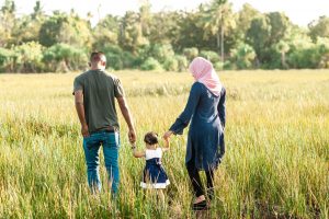 Family of four walking hand-in-hand through a sunlit, tall-grass field with trees in the background.