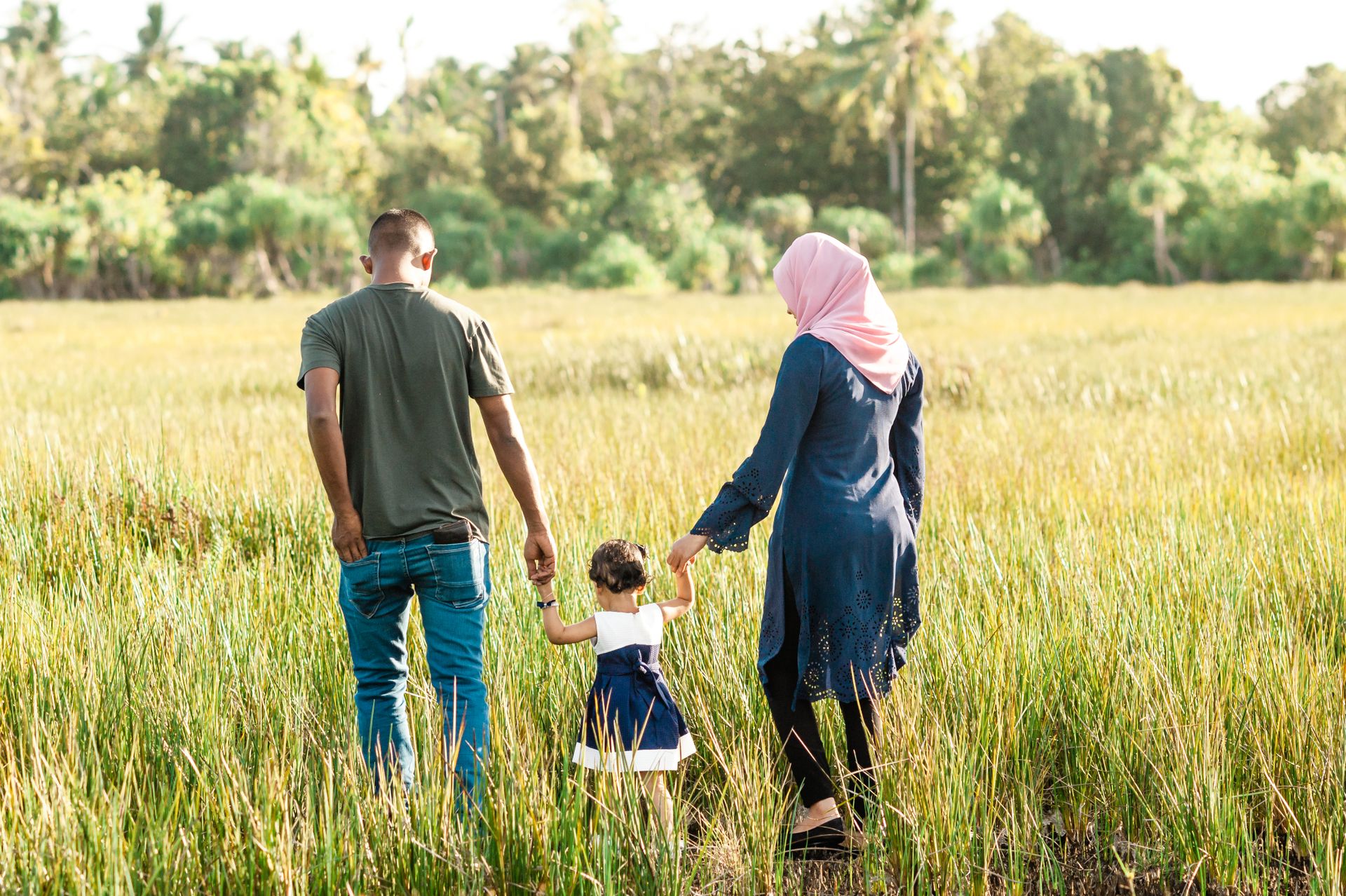 Family of four walking hand-in-hand through a sunlit, tall-grass field with trees in the background.