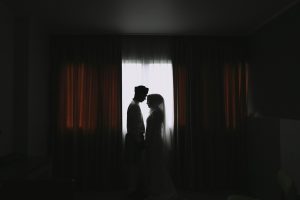 Silhouetted bride and groom facing each other in a dim room, backlit by a bright window framed by curtains.
