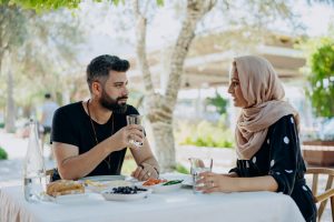 A man and woman wearing hijab sit at an outdoor cafe table, sharing a meal and conversation under trees.
