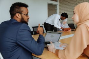 Diverse coworkers collaborate at a conference table; a man leans over to help a woman with a tablet.