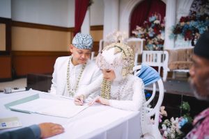 Bride and groom in traditional white attire sign a marriage document at a ceremony, ornate headpieces and floral backdrop behind them.